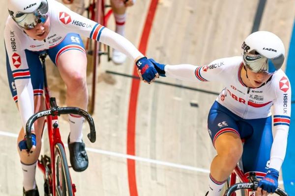 Two female cyclists on a track cycle side by side, gripping each others' hands
