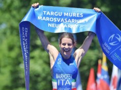 Triathlete Sandra Maihofer smiles as she crosses the finish line with a blue finish tape held aloft in victory