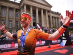 An athete wearing orange has his hands in the air and a medal around his neck. In the background is the red finish carpet and Bolton Albert Halls.