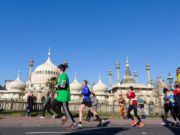 Colourful runners pass an ornate white building topped with domes. The sky behind is a bright blue.