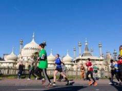 Colourful runners pass an ornate white building topped with domes. The sky behind is a bright blue.