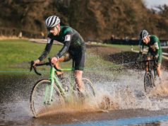 Two male cyclists ride through a large puddle - water sprays up high.