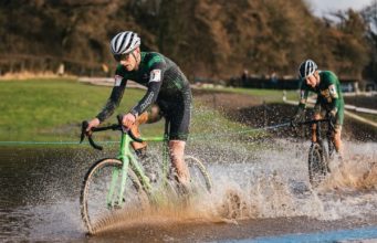 Two male cyclists ride through a large puddle - water sprays up high.
