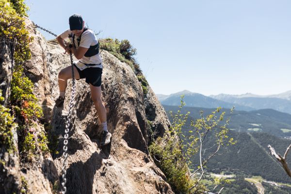 Photo of a runner scaling a high up rock face, with the aid of an iron chain.