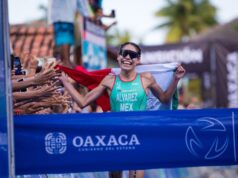 Female triathlete in green vest and sunglasses wearing the Mexican flag draped over her shoulders approaches the blue finish tape.