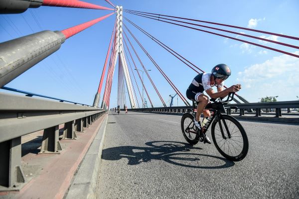 A cyclist rides over a bridge. His head is down and he holds his racing bars.