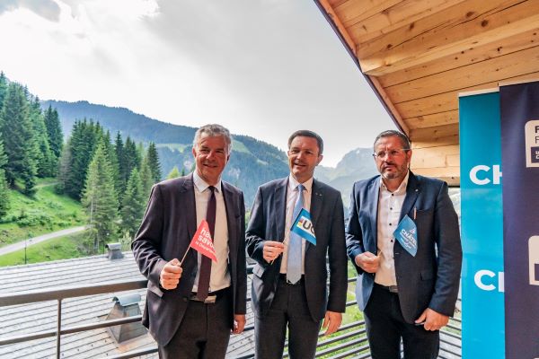 Three men in suits wave small flags on a wooden balcony, with mountains behind them.