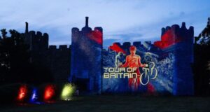 A castle at dusk lit up in red and blue with an image of a cyclist and the words 'Tour of Britain'