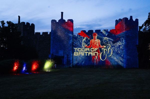 A castle at dusk lit up in red and blue with an image of a cyclist and the words 'Tour of Britain'