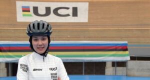 Female athlete stands in cycling gear with arms folded, smiling at the camera. A UCI banner is in the background.
