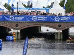 A photo of athletes swimming underneath a bridge which has thousands of spectators watching