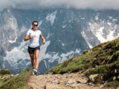 A femal runner wearing white t-shirt, black shorts, sunglasses and a running watch runs with snowy mountains behind her.