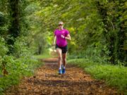 Pregnant athlete wearing pink t-shirt, black shorts and sunglasses run along a woodland trail towards the camera.