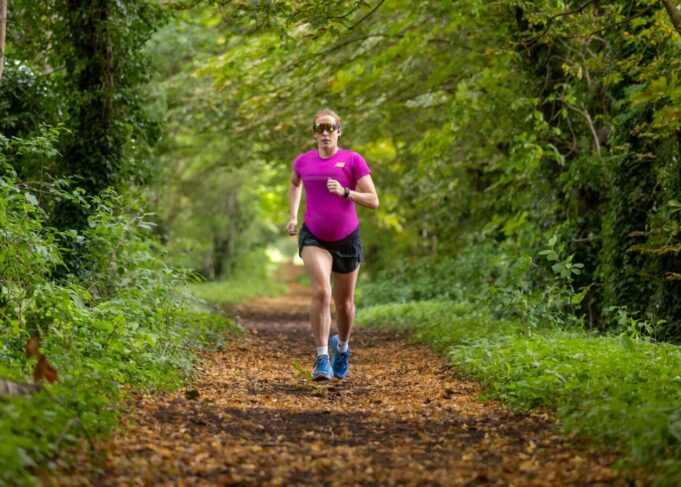 Pregnant athlete wearing pink t-shirt, black shorts and sunglasses run along a woodland trail towards the camera.