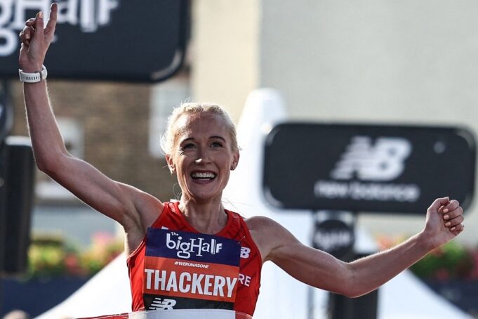 Female athlete smiles and holds her arms high. She is blonde, and is wearing a red top. Her race number says The Big Half, and her surname in capital letters, THACKERY