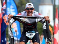 Male triathlete bends towards the camera clutching an IRONMAN finish tape in his hands. He looks to be yelling in victory.