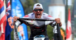Male triathlete bends towards the camera clutching an IRONMAN finish tape in his hands. He looks to be yelling in victory.