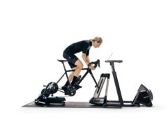 Female athlete stands up atop a machine emulating a bike in front of a screen.
