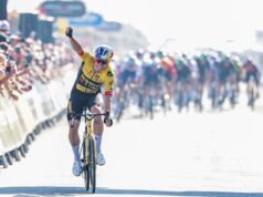 Wout van Aert in a yellow jersey holds one arm in the air in victory as he takes the stage win. The rest of the cyclists are visible in the background.