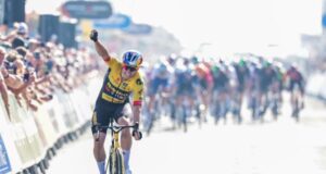 Wout van Aert in a yellow jersey holds one arm in the air in victory as he takes the stage win. The rest of the cyclists are visible in the background.