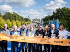 7 men and 1 woman hold a banner that reads Superhalfs welcomes Berlin. Behind them the Brandenburg Gate is visible, and a road that is full of runners.