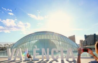 A lady reclines on the letter L of a large sign saying Valencia in front of a glass dome. Somebody in the foreground is taking her photograph on a red smartphone.