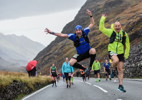 Runners mostly walk up one of the hills. In the foreground, a man in a blue T-shirt leaps into the air for the camera man, and a man in a yellow jacket next to him waves. The weather looks drizzly, but the landscape is beautiful and mountainous.