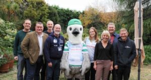 A group of 8 people in business wear stand either side of somebody dressed up as a giant seagull wearing ed trainers, sports kit and a green cap.