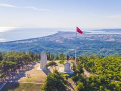 A white war memorial stands atop a hillside overlooking the coast. The climate looks warm.