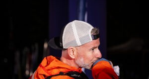 Scott wears an orange jacket, race pack and a white cap worn backwards. He's looking off camera, and appears focused. It's night time.