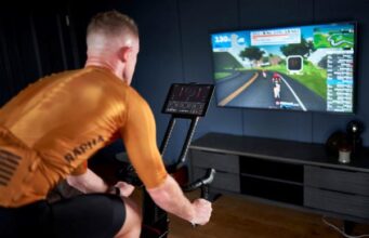 man wearing orange cycling jersey sits on a Wattbike. In front of him on a large TV screen is a race graphic of a road that he is virtually cycling along.