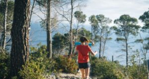 A female runner runs away from the camera down a rocky, tree-lined path, towards the coast.