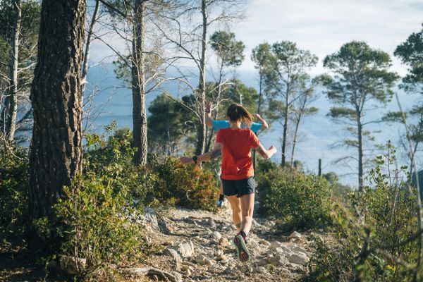 A female runner runs away from the camera down a rocky, tree-lined path, towards the coast.