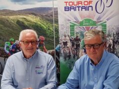 Two elderly gentlemen pose in front of a screen showing an image of the tour of Britain.