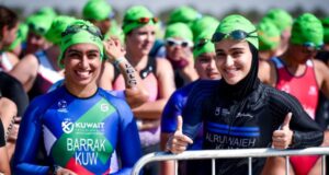 A group of female triathletes, all wearing green swim hats, wait to start their swim. Two women at the front smile at the camera.