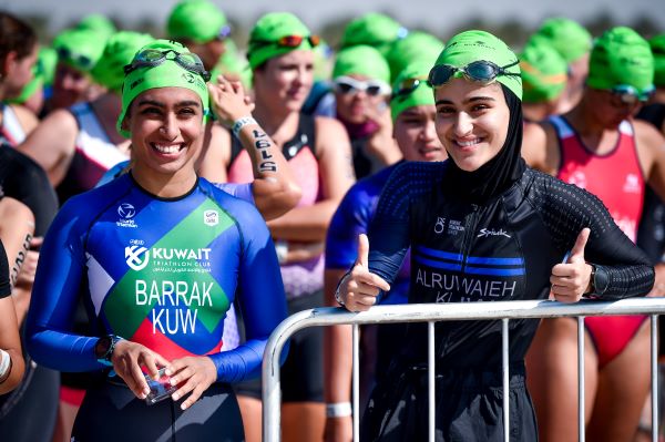 A group of female triathletes, all wearing green swim hats, wait to start their swim. Two women at the front smile at the camera.