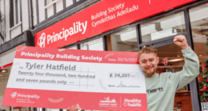 A young man stands outside a building society holding a huge cheque worth £24,207