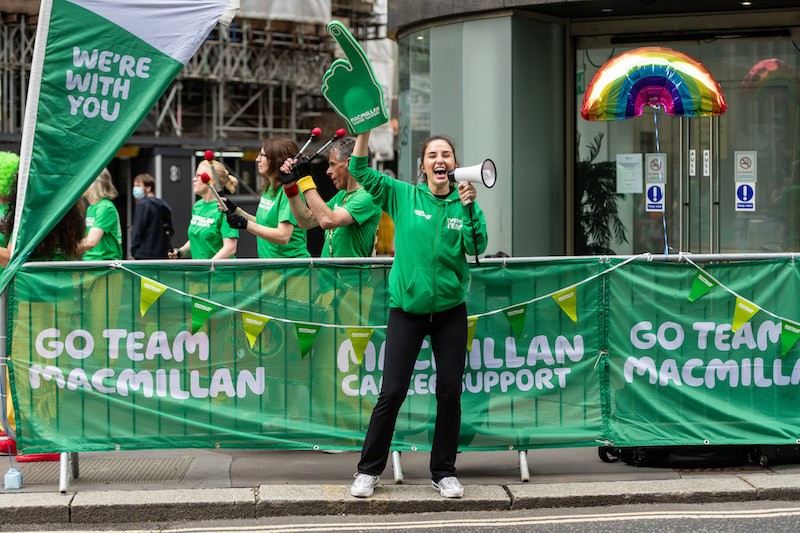 Macmillan cheer point at a running event. A ladt has a mega phone in one hand and a large green foam hand on the other pointing up
