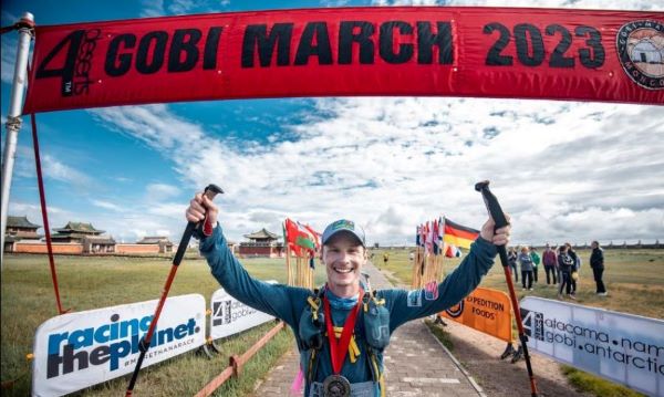 Man smiles and holds poles aloft in victory as he poses under finishing banner of Gobi March 2023