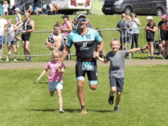Smiling man in tri suit runs towards camera, holding the hands of two children, a girl and a boy.