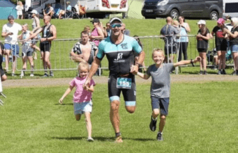 Smiling man in tri suit runs towards camera, holding the hands of two children, a girl and a boy.
