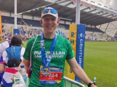 A man stands in a sports stadium wearing a green Macmillan T-shirt, white cap with sunglasses pushed onto the peak. He is smiling and wearing a medal around his neck.