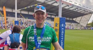 A man stands in a sports stadium wearing a green Macmillan T-shirt, white cap with sunglasses pushed onto the peak. He is smiling and wearing a medal around his neck.