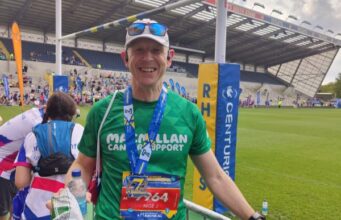 A man stands in a sports stadium wearing a green Macmillan T-shirt, white cap with sunglasses pushed onto the peak. He is smiling and wearing a medal around his neck.