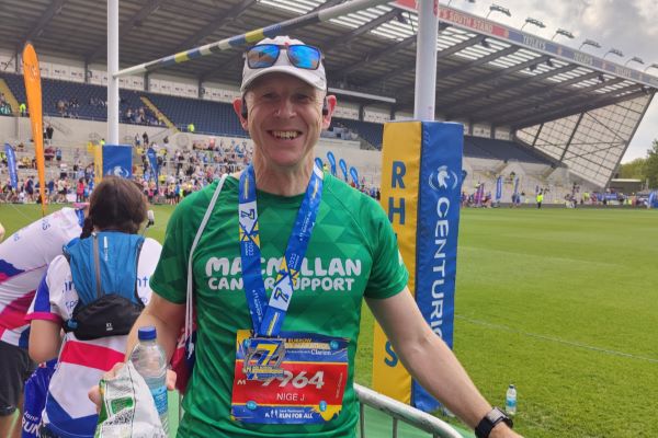 A man stands in a sports stadium wearing a green Macmillan T-shirt, white cap with sunglasses pushed onto the peak. He is smiling and wearing a medal around his neck.