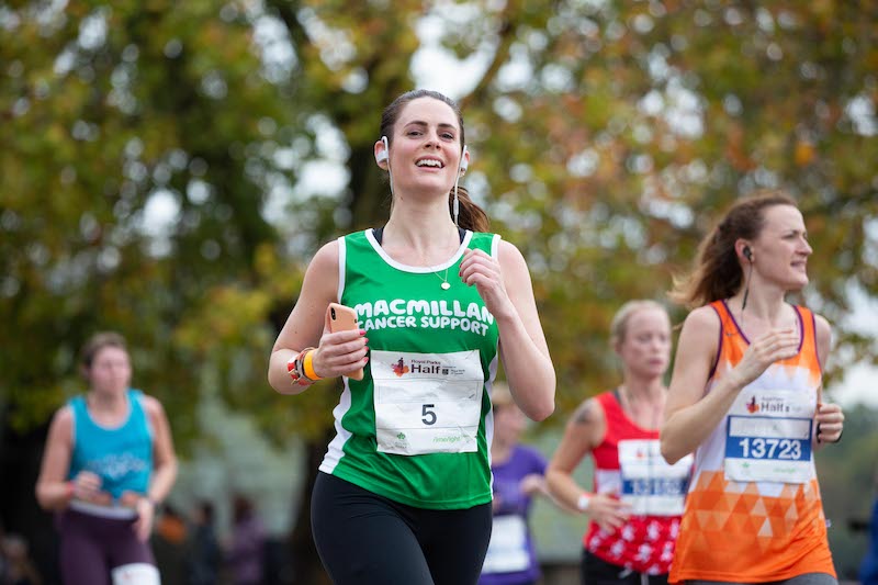 Royal Parks A brown haired lady smiling whilst running represting Macmillan. Wearing the green vest and her racing bib number with a number 5 on it.