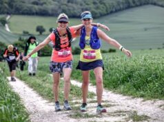 Two women in colourful running kit with their arms around each other, smile at the camera. They are on a countryside track.