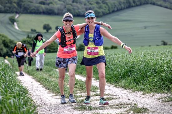 Two women in colourful running kit with their arms around each other, smile at the camera. They are on a countryside track.