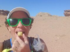 Woman with short black hair, wearing green sunglasses and white cap, stands in desert eating a cake.