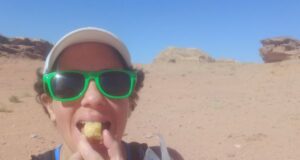 Woman with short black hair, wearing green sunglasses and white cap, stands in desert eating a cake.